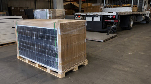 Tarima de madera llena de paneles solares en cajas con film en un piso de almacén con un camión de plataforma hidráulica detrás