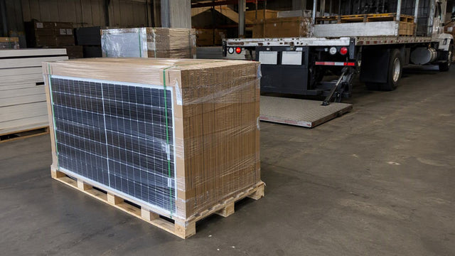 Tarima de madera llena de paneles solares en cajas con film en un piso de almacén con un camión de plataforma hidráulica detrás