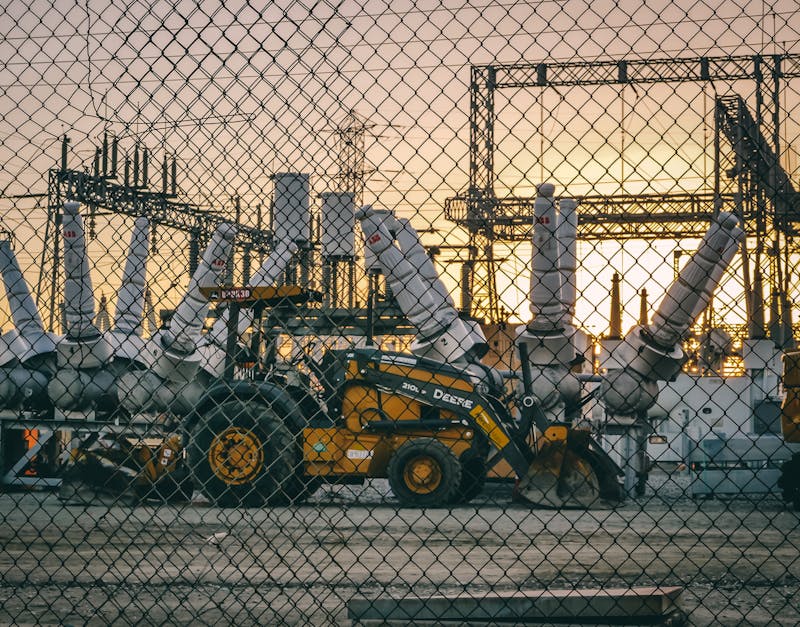 Yellow construction vehicle at industrial substation behind chain link fence at sunset