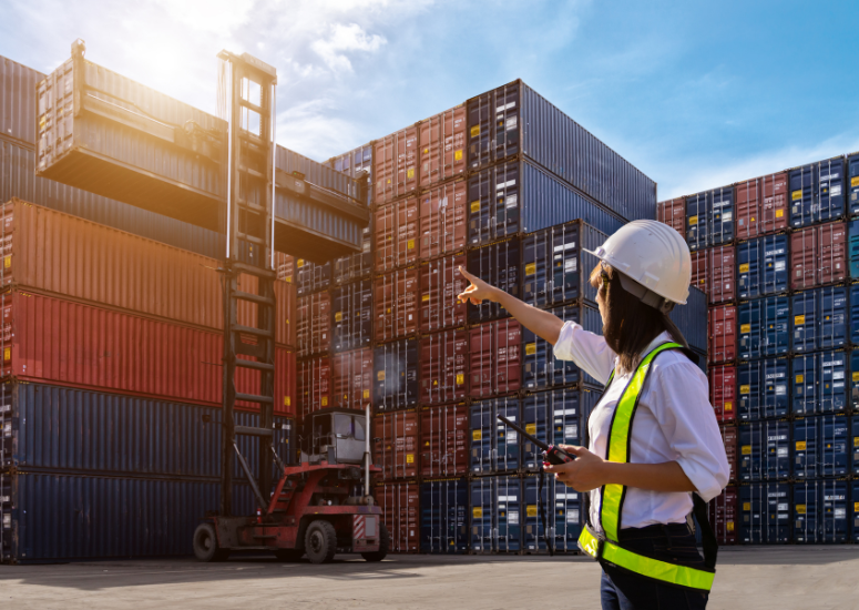 Person in a hard hat and safety vest pointing at stacked shipping containers with a forklift in the background.