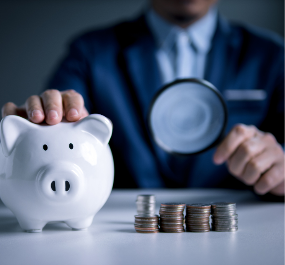 Person examining coins with a magnifying glass next to a piggy bank on a table.