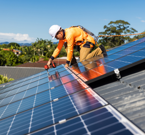 Worker installing solar panels on a roof with a clear sky and trees in the background