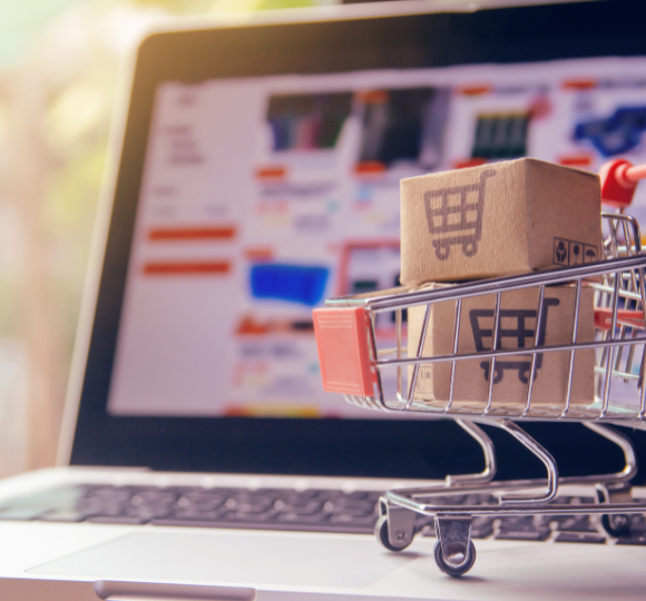 Small shopping cart with boxes in front of a laptop displaying an e-commerce website.